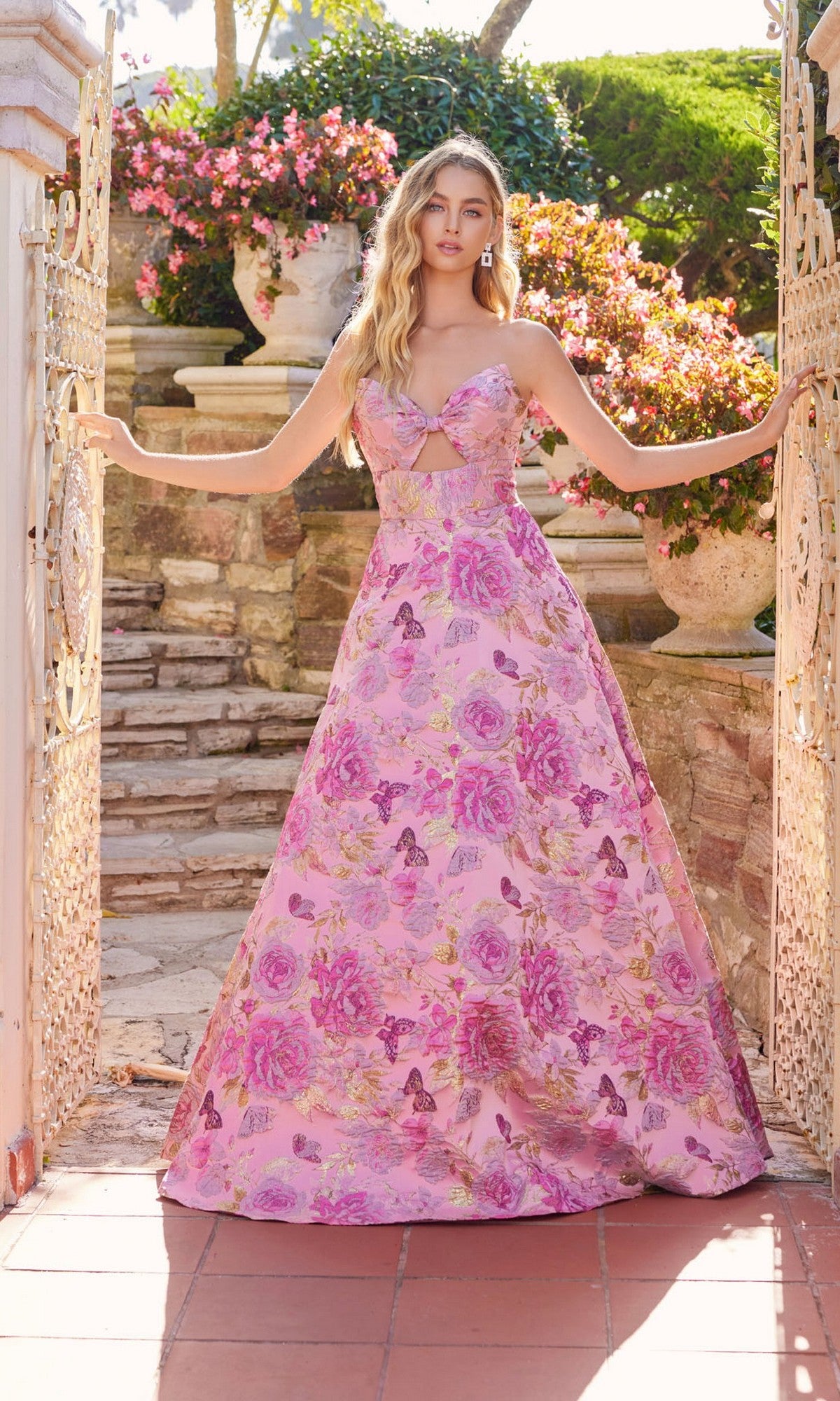Wearing the Juliet JT2735H long prom dress, a woman stands by an open wrought iron gate, stone steps and blooming flowers behind her.
